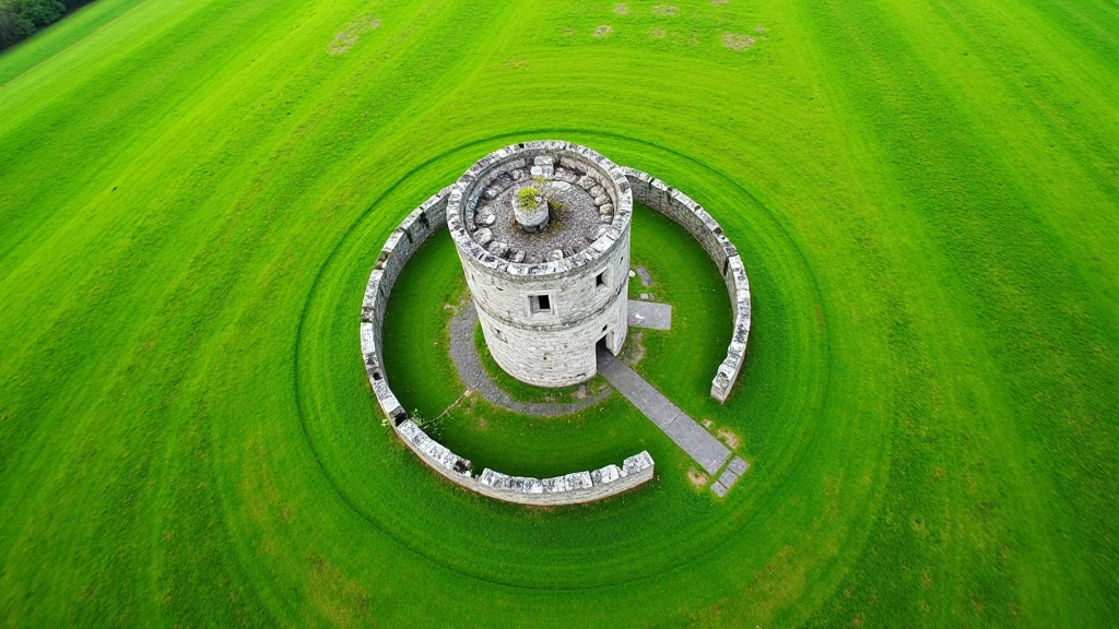 Aerial view of round tower and stone monastic buildings scattered across green Irish landscape with stone walls